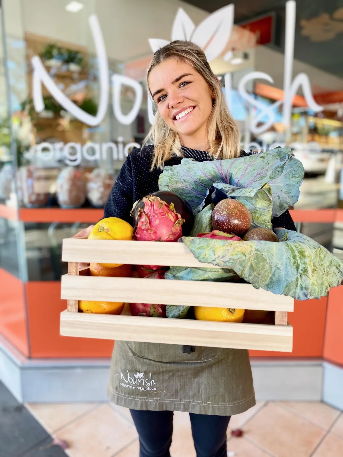 A Nourish team member smiling outside the store, holding a crate of organic produce